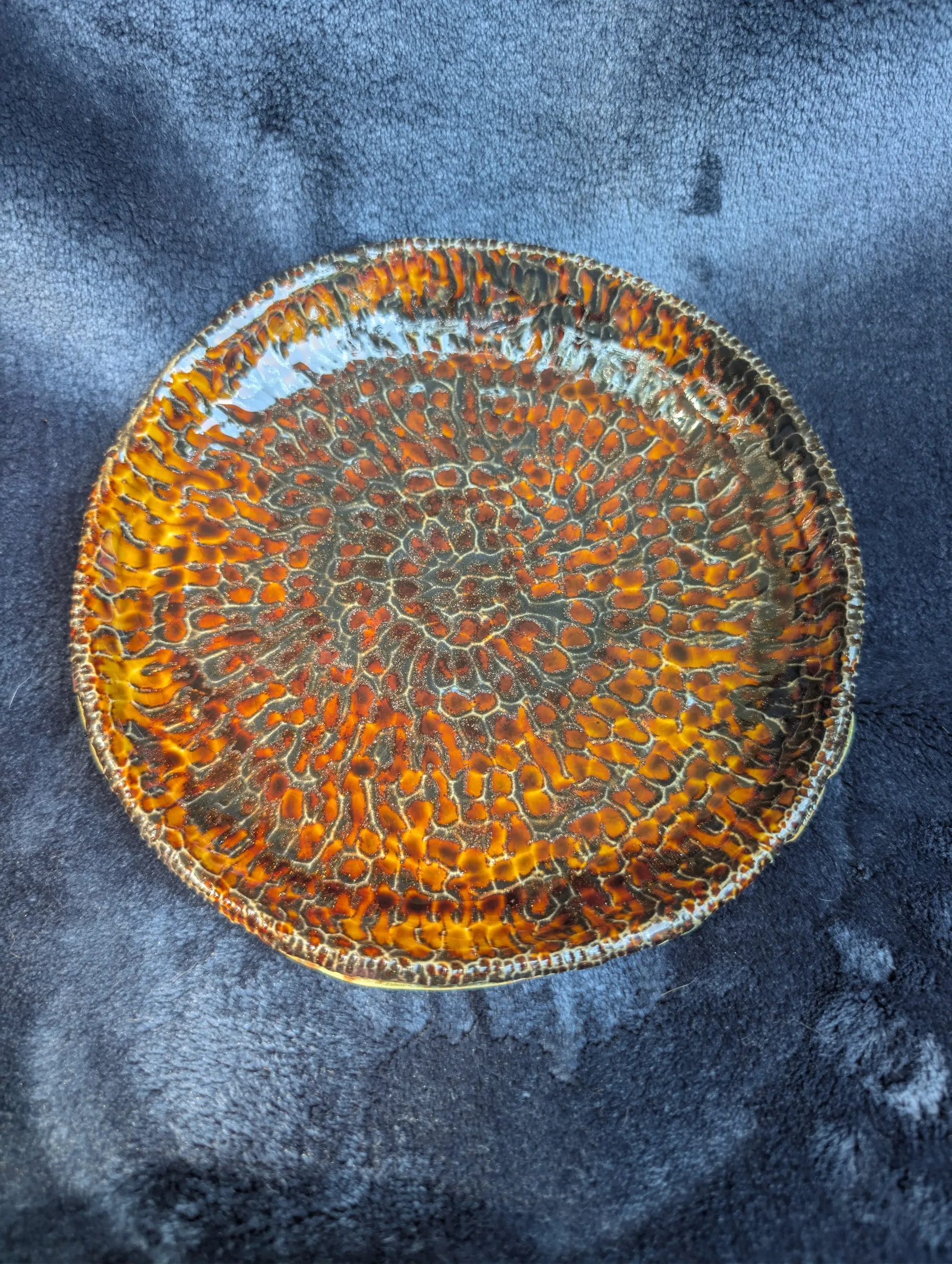 A round plate covered edge to edge in a glossy amber and orange glaze patterned with fine crackle, photographed on a blue cloth.