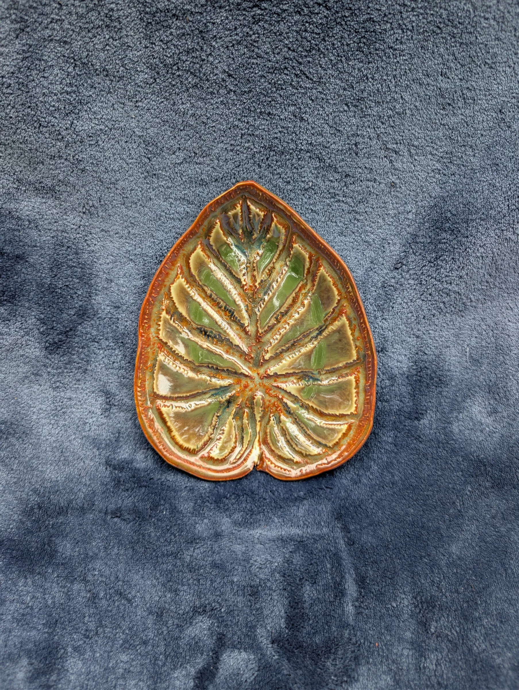 A pointed, arrow-shaped glazed dish with a green well, an orange-brown rim and radiating impressed veins, on a blue cloth.