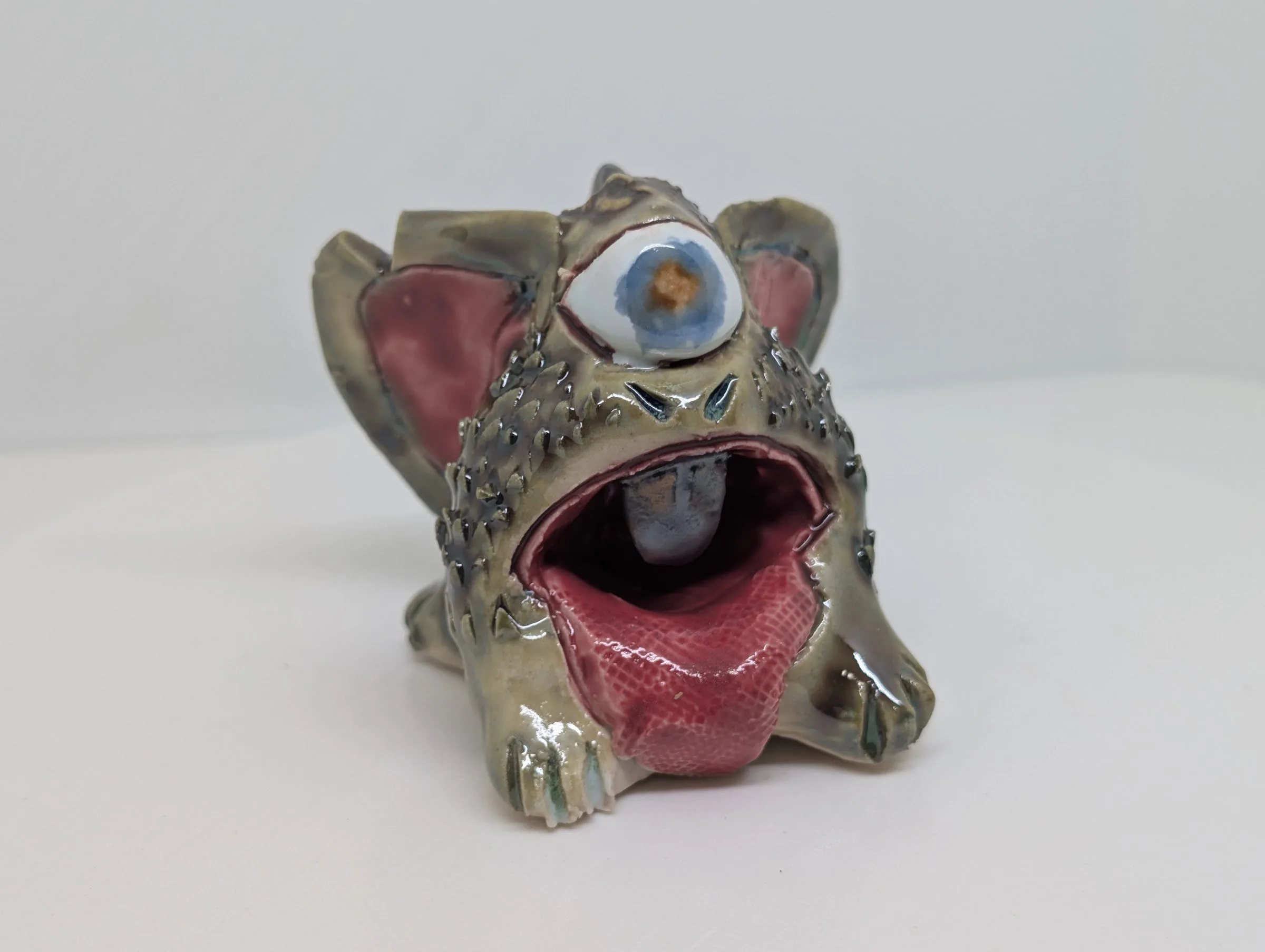 A glazed ceramic creature with a wide open mouth, a pink tongue, one large eye and small pointed ears, photographed on a plain white surface.