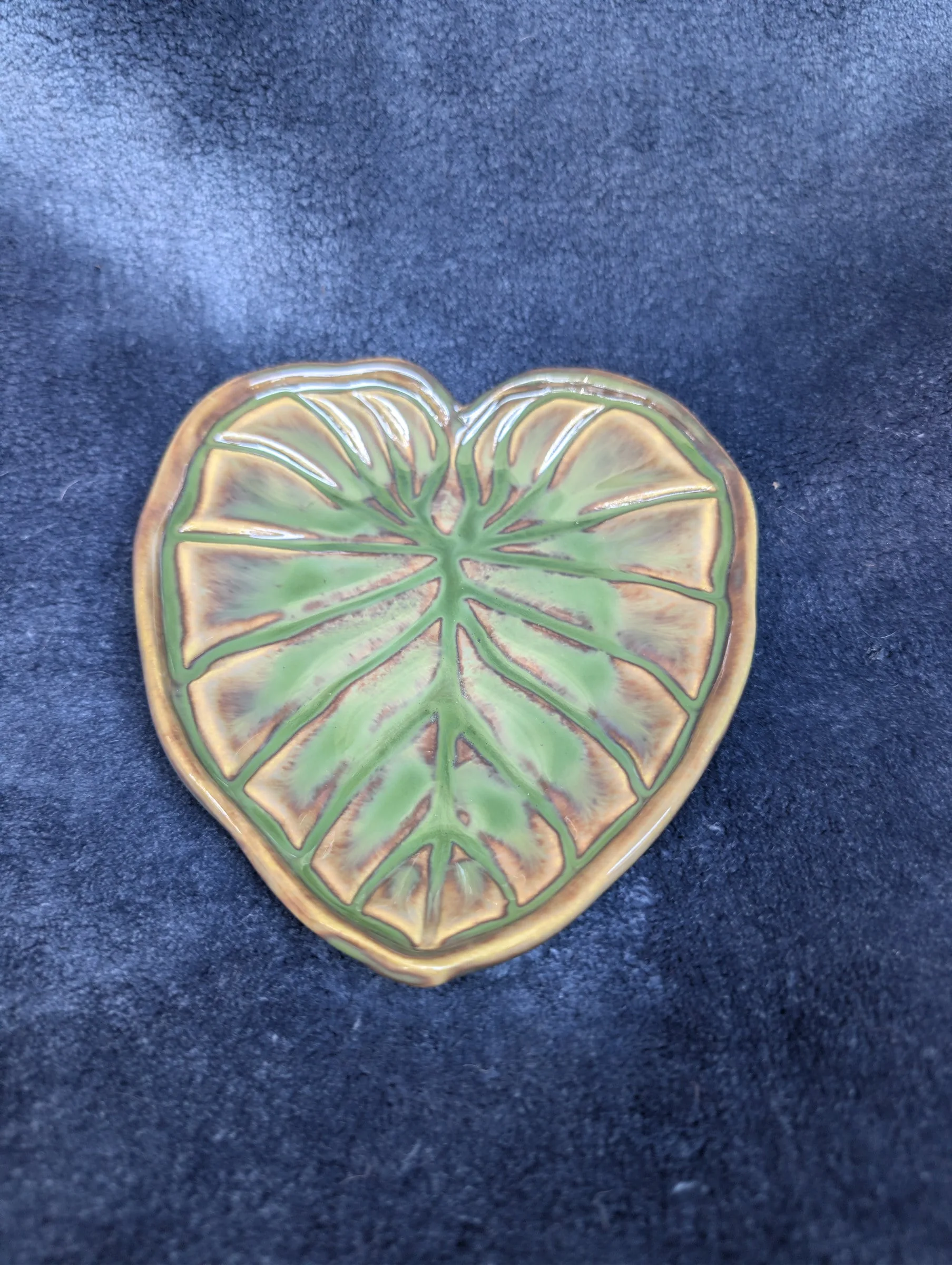 A shallow glazed dish shaped like a heart-shaped leaf, with a green body, a thick amber rim and radiating veins, photographed on a dark blue cloth.