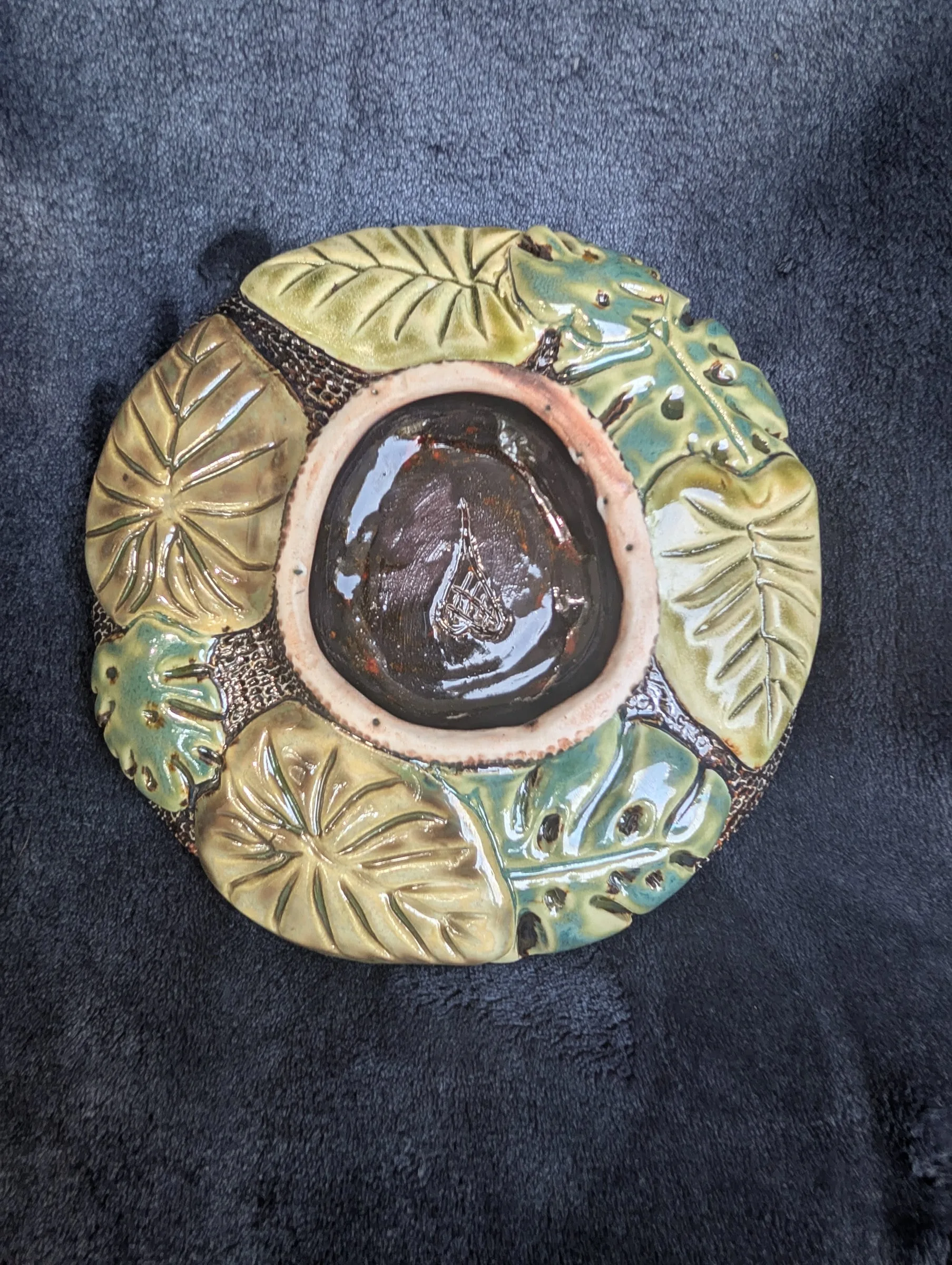 A round plate with pale green glazed leaves arranged in a ring around a deep brown glossy center, each leaf outlined by a raised edge.