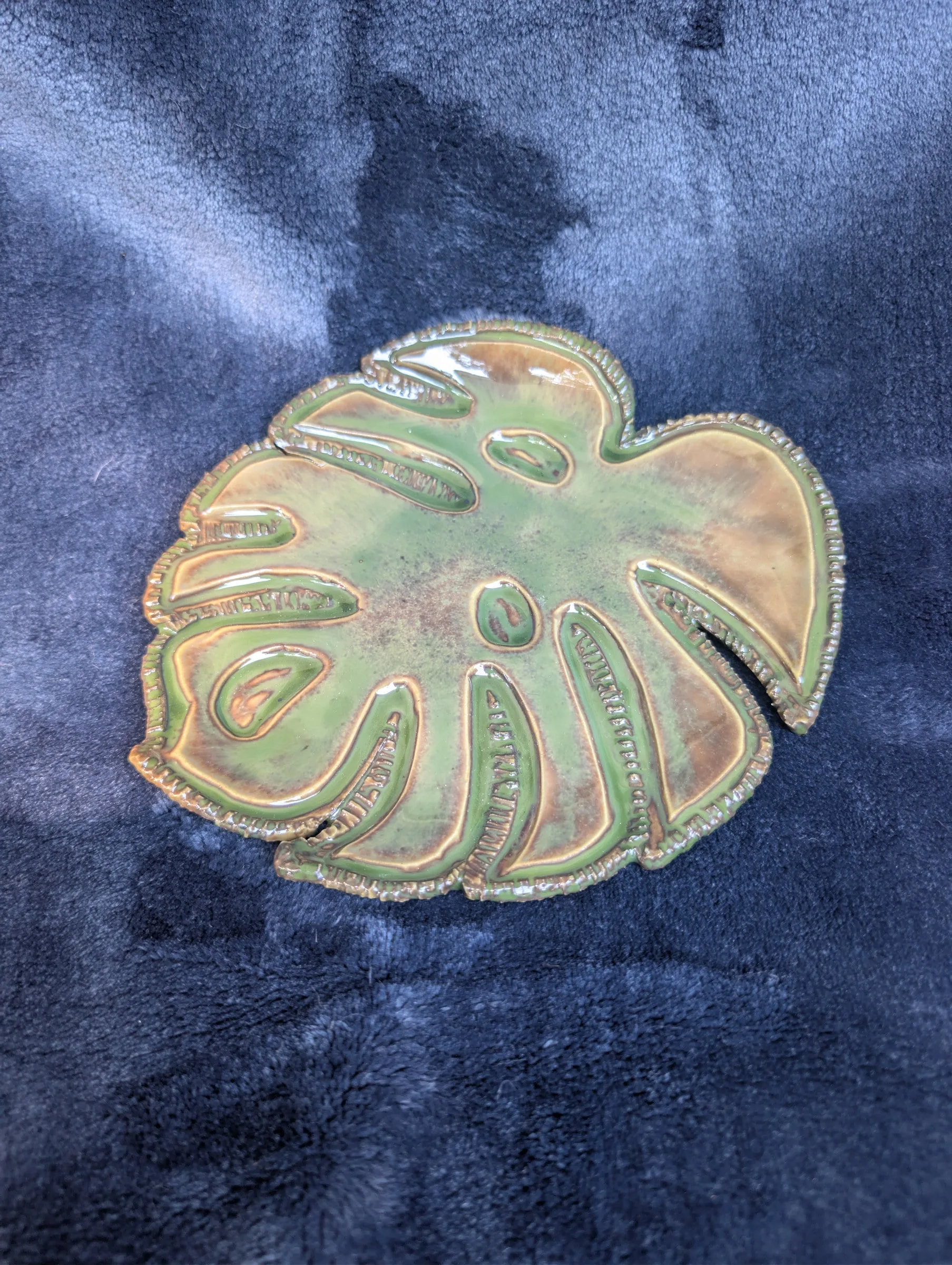 A wide, low platter in the shape of a monstera leaf with several cut-out windows, a green and amber rim and a paler mottled center, on a blue cloth.