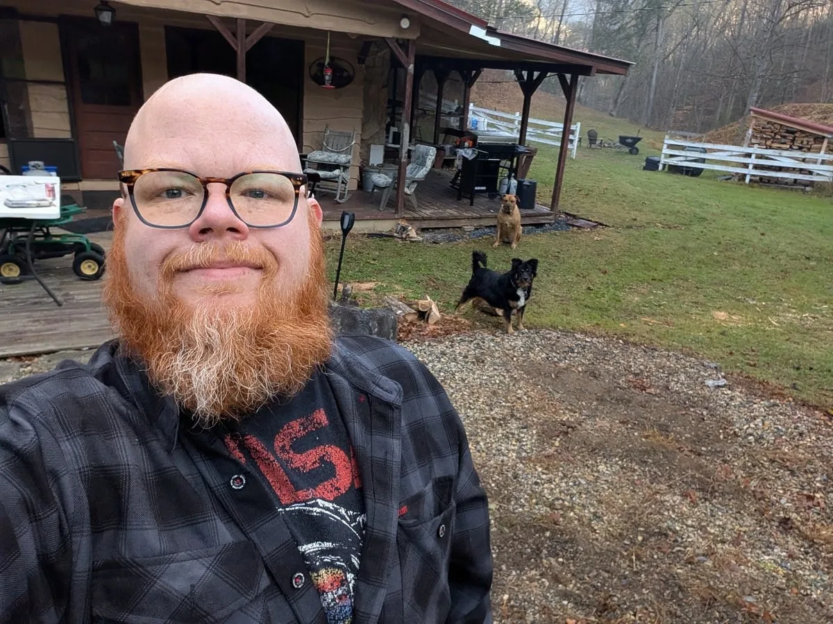 Casey Hartnett outdoors on the studio property, with a red beard, glasses and a plaid shirt, and two dogs behind him.